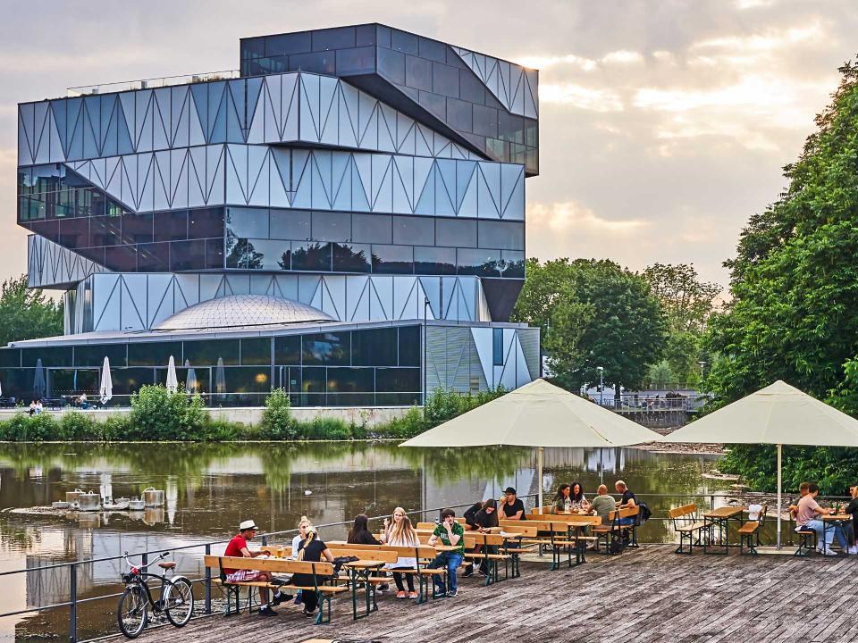 Gäste sitzen gemütlich an Tischen auf einer Holzterrasse am Flussufer, im Hintergrund ein markantes, modernes Glasgebäude____