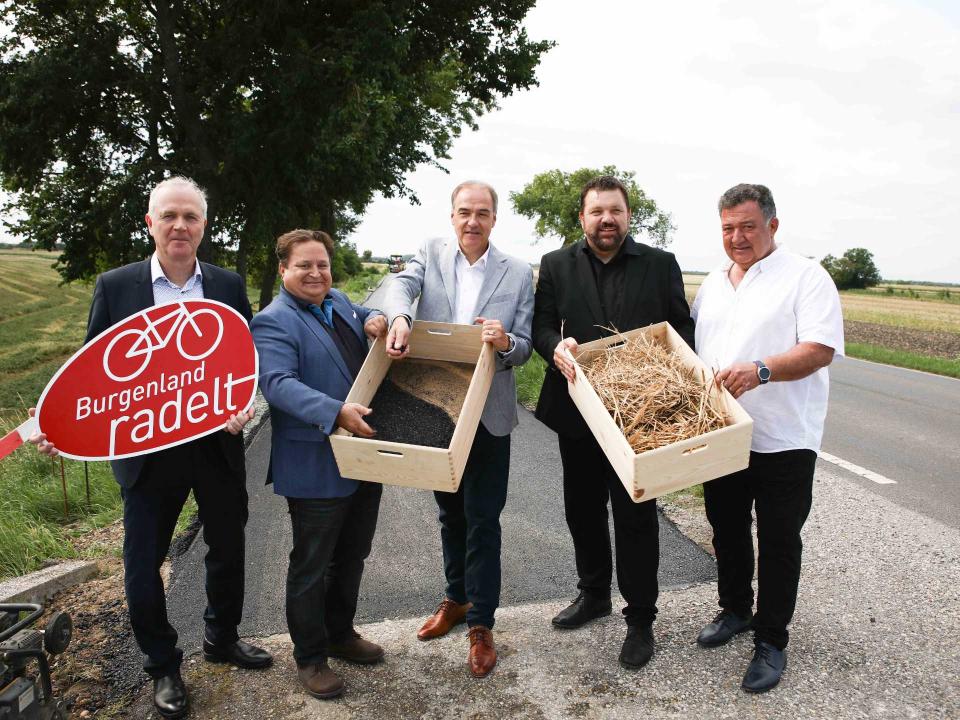 Fünf Männer stehen mit Kisten voller Saatgut und Stroh neben einem „Burgenland radelt“-Schild auf Landstraße____