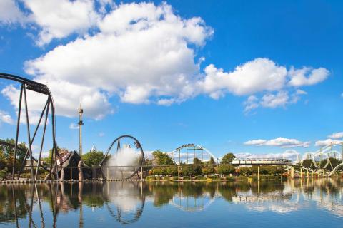 Achterbahnen und Wasserattraktion spiegeln sich im See unter blauem Himmel im Freizeitpark