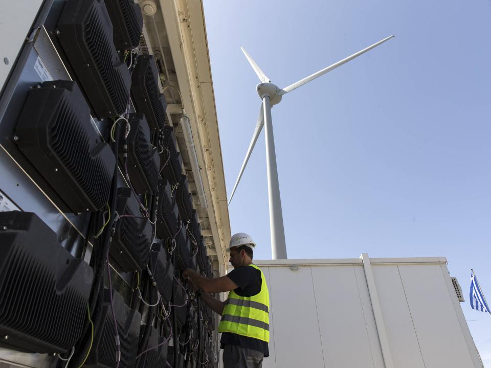 Techniker mit Schutzhelm und Warnweste arbeitet an Energiekomponenten, dahinter Windturbine und blauer Himmel____