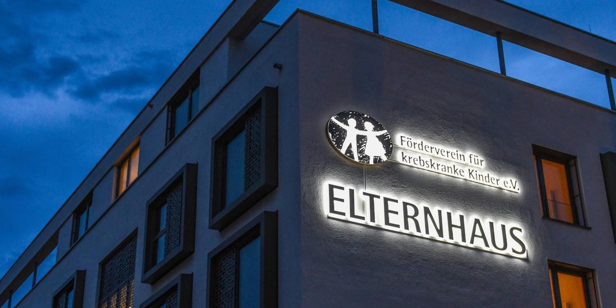 Haus bei Dunkelheit mit Logo. Text: ELTERNHAUS. Förderverein für krebskranke Kinder e.V. Das beleuchtete Logo zeigt ein Kind und einen Erwachsenen