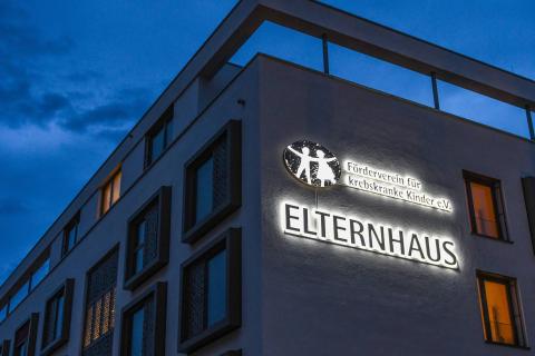 Haus bei Dunkelheit mit Logo. Text: ELTERNHAUS. Förderverein für krebskranke Kinder e.V. Das beleuchtete Logo zeigt ein Kind und einen Erwachsenen