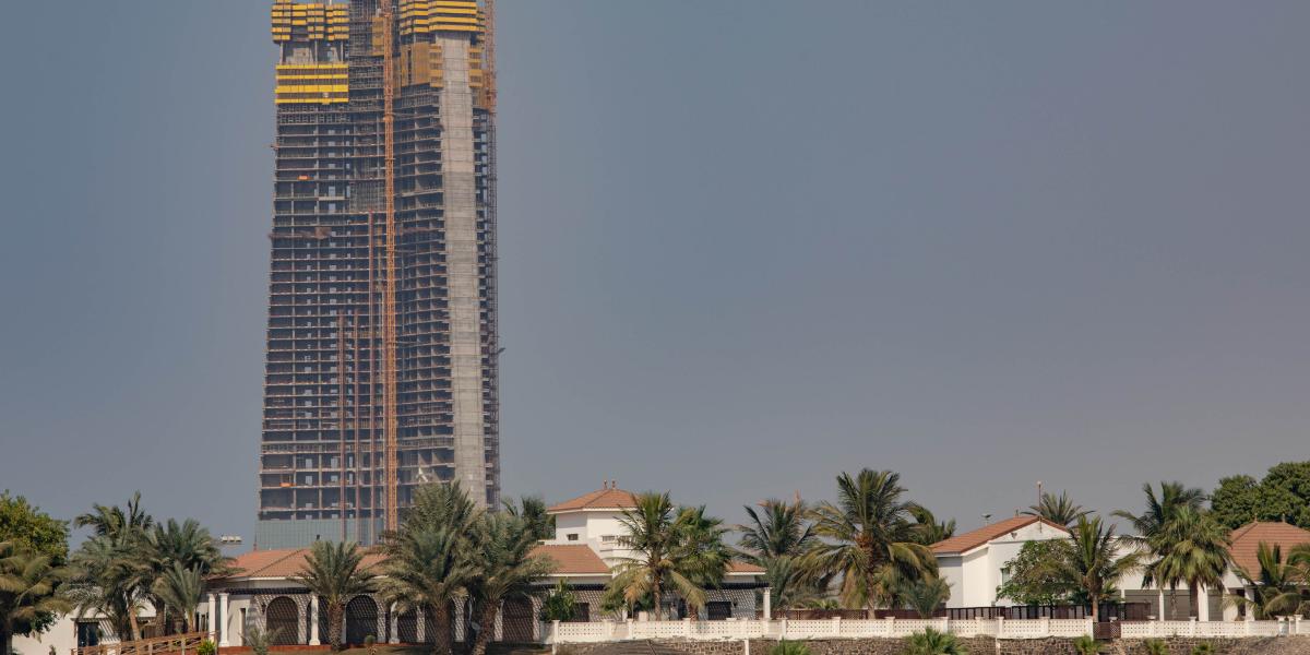 Ein Hochhaus im Bau mit gelben Sicherheitsabdeckungen und Baukränen, vor niedrigen Häusern und Palmen bei klarem Himmel