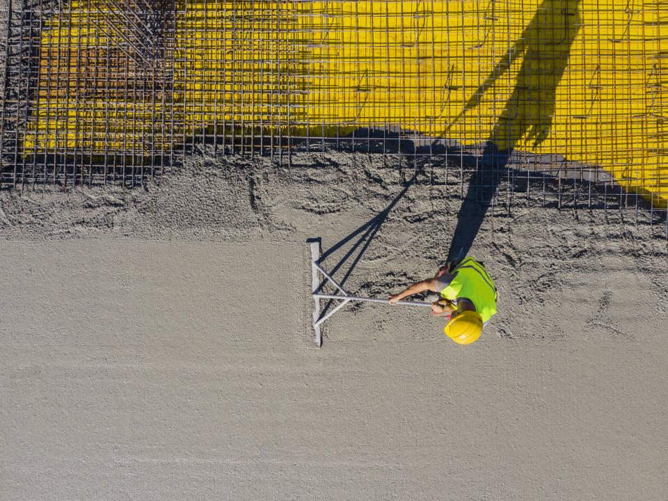 Bauarbeiter mit Helm und Weste glättet frischen Beton neben Armierungseisen auf Baustelle.____