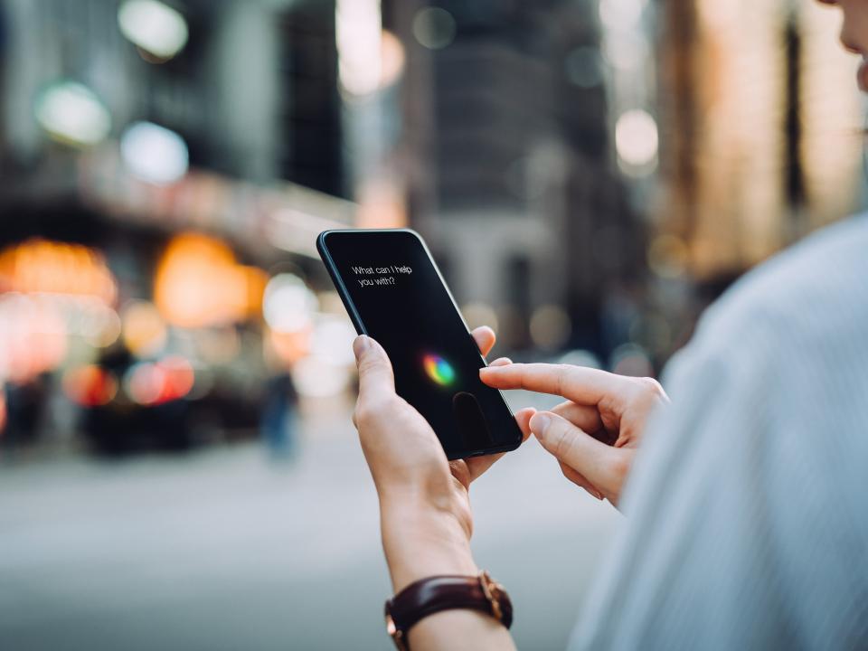 Person nutzt Sprachassistent auf Smartphone in Großstadt, Text „What can I help you with?“ auf Display sichtbar.____