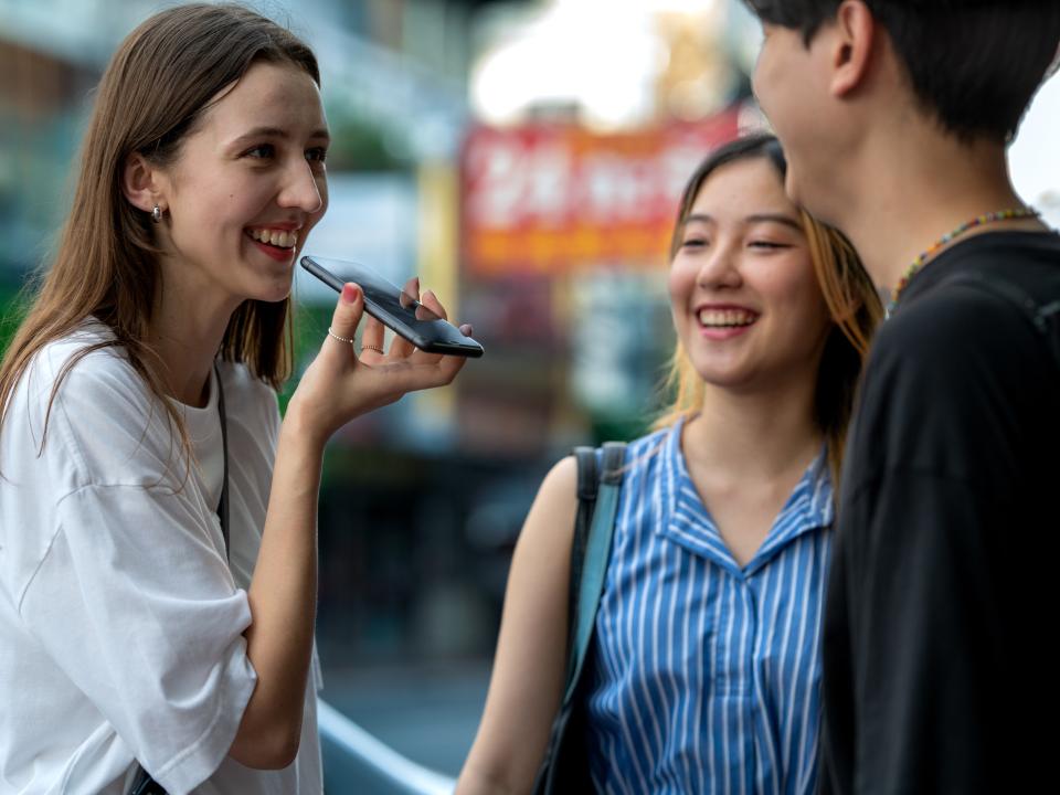 Junge Frau spricht fröhlich in Smartphone-Sprachsteuerung, zwei Freund:innen stehen daneben und lachen.____