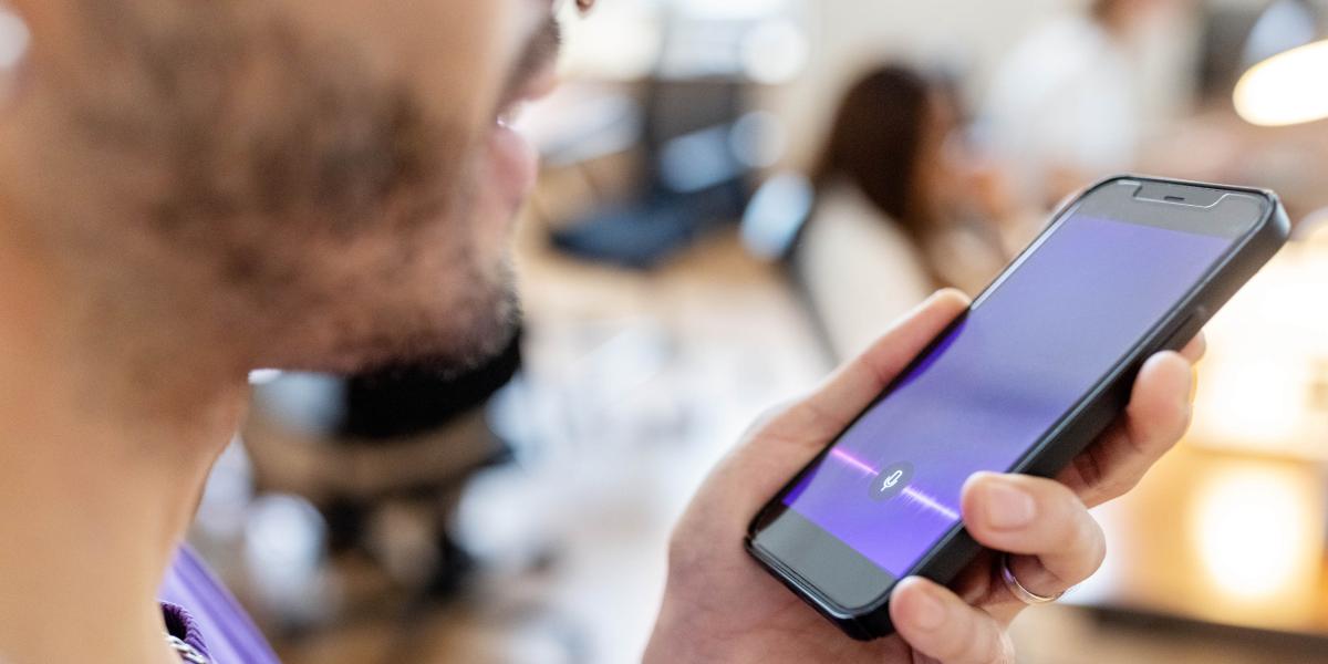 Mann spricht in Smartphone mit aktivierter Sprach-KI-App, Fokus auf Display mit bunter Audio-Wellenanzeige.