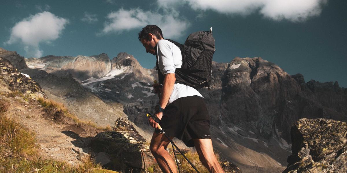 Mann wandert mit Trekkingstöcken einen felsigen Bergpfad hinauf, hohe Berge im Hintergrund.