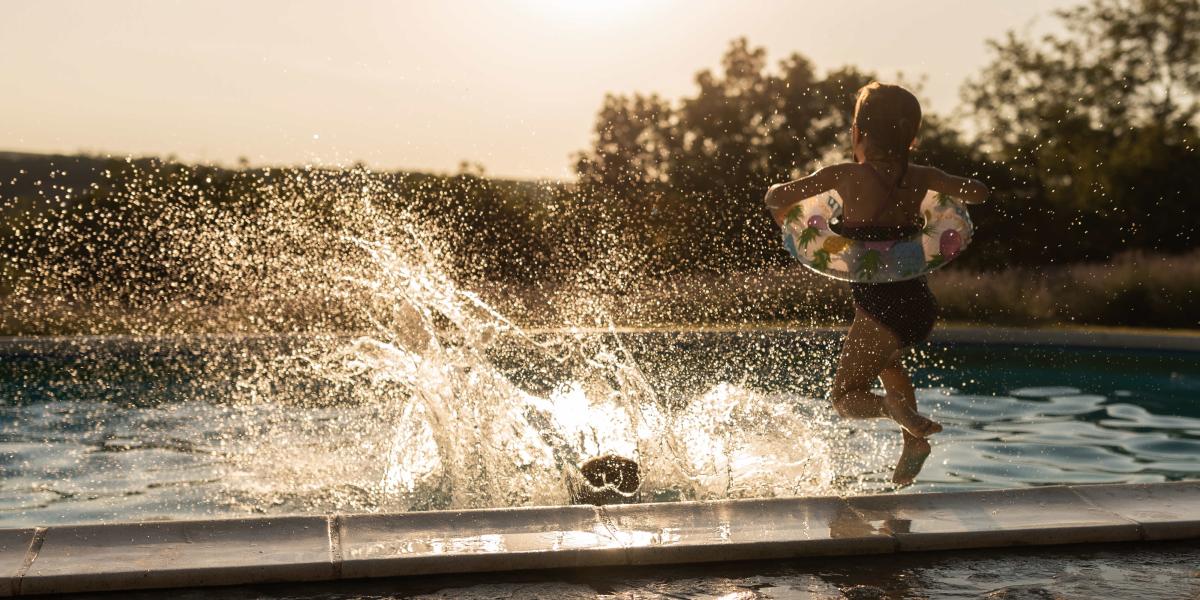 Kleines Kind mit Schwimmring springt in einen Pool, Wasser spritzt in der Abendsonne auf.
