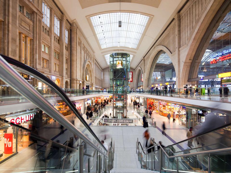 Leipziger Hauptbahnhof mit Rolltreppen, Geschäften und Glasfassade, belebt mit Reisenden und Besuchern____