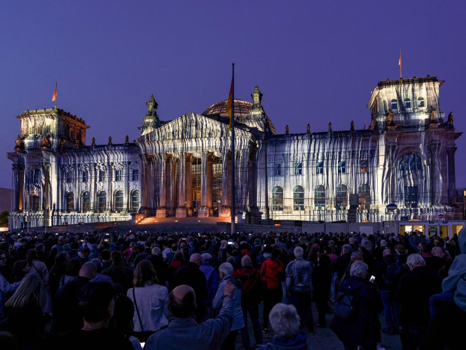 Beleuchteter Reichstag in Berlin mit Projektionen des verhüllten Gebäudes, umgeben von Menschen bei Nacht____