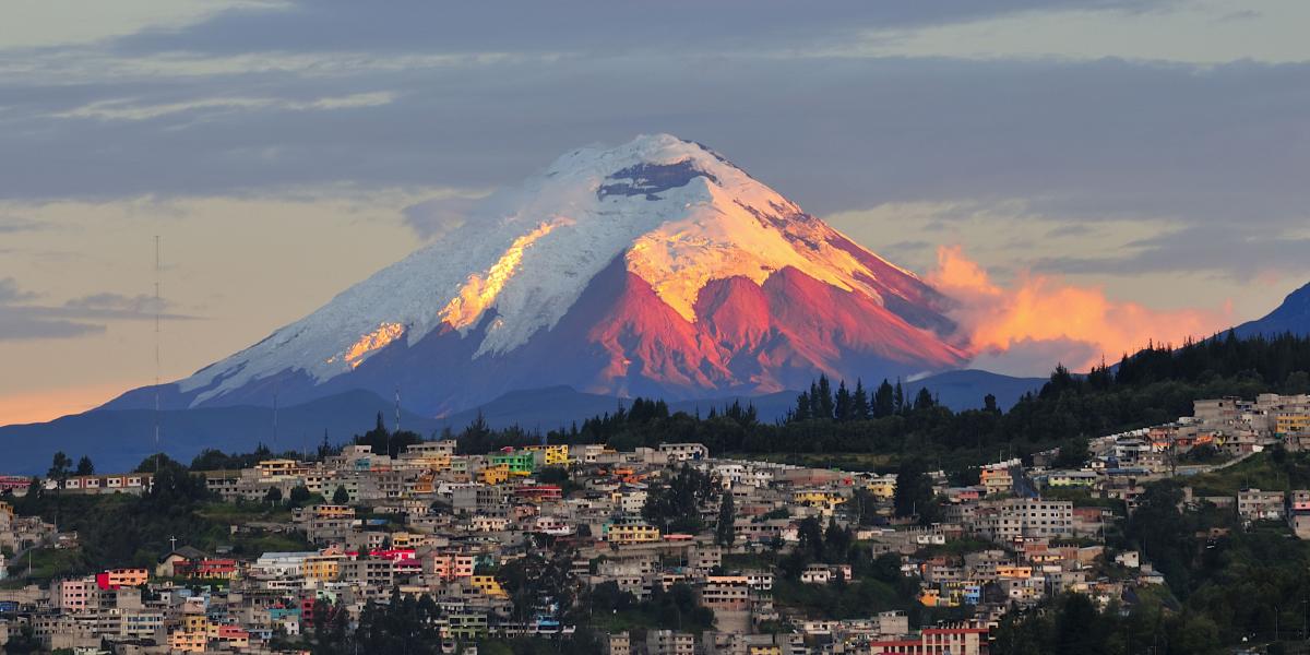 Vulkan Cotopaxi im Sonnenuntergang, umgeben von Stadt und bunten Häusern.