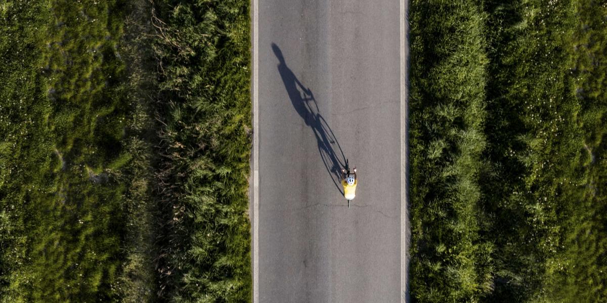 Luftaufnahme eines allein fahrenden Radfahrers mit gelbem Trikot auf gerader Straße durch grüne Landschaft