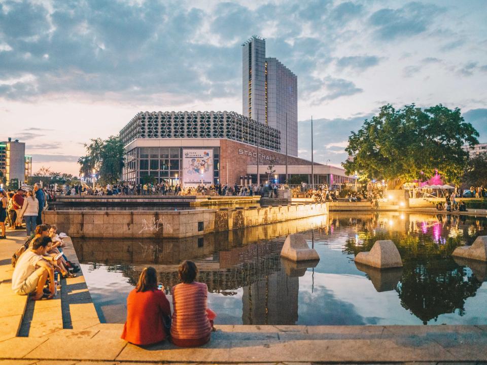 Stadtplatz in Chemnitz mit Teich und Sitzbereichen, belebt mit Menschen, Stadthalle und Hochhaus im Hintergrund bei Abendstimmung____