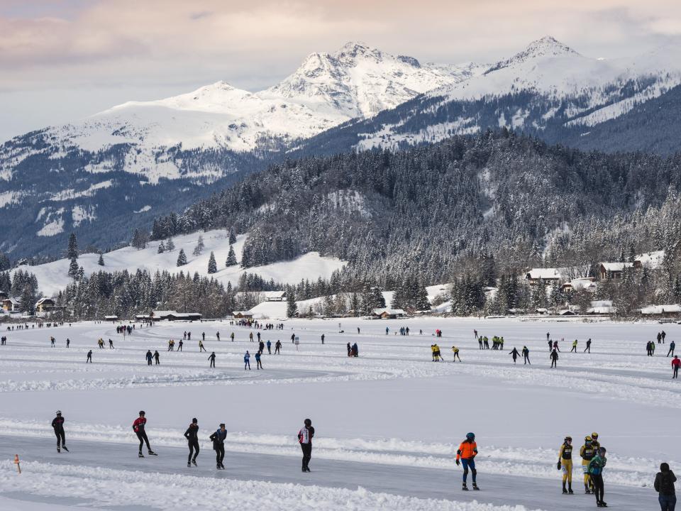 Zahlreiche Schlittschuhläufer auf einer weitläufigen Eisfläche in schneebedeckter Berglandschaft.____