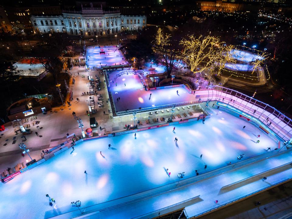 Nachtaufnahme einer beleuchteten Eislaufbahn mit Schlittschuhläufern, umgeben von dekorierten Bäumen.____