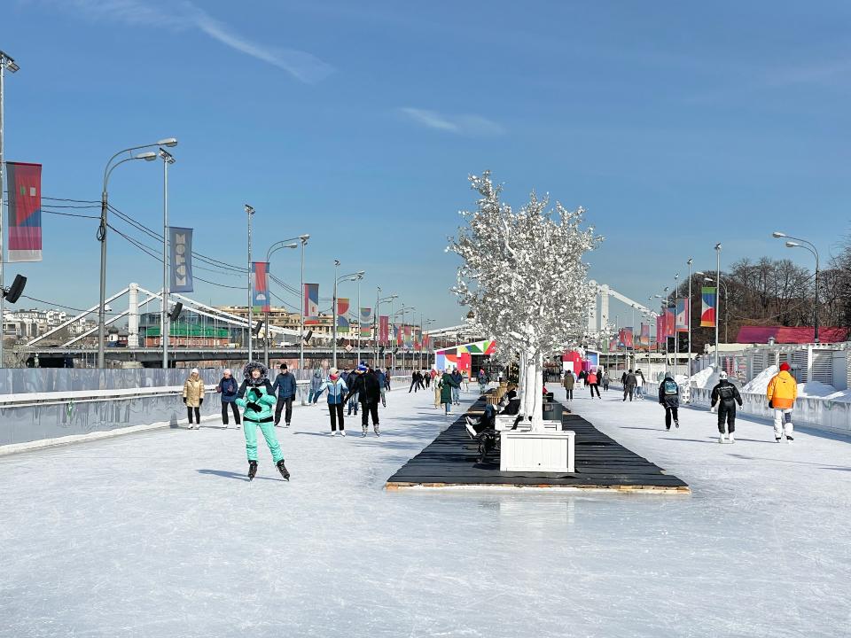 Schlittschuhläufer auf einer breiten Eisbahn bei Sonnenschein, mit Dekobäumen und Stadtfahnen im Hintergrund.____