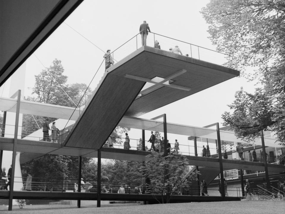 Futuristischer Ausstellungspavillon mit erhöhter Plattform und vielen Besuchern auf mehreren Ebenen.____