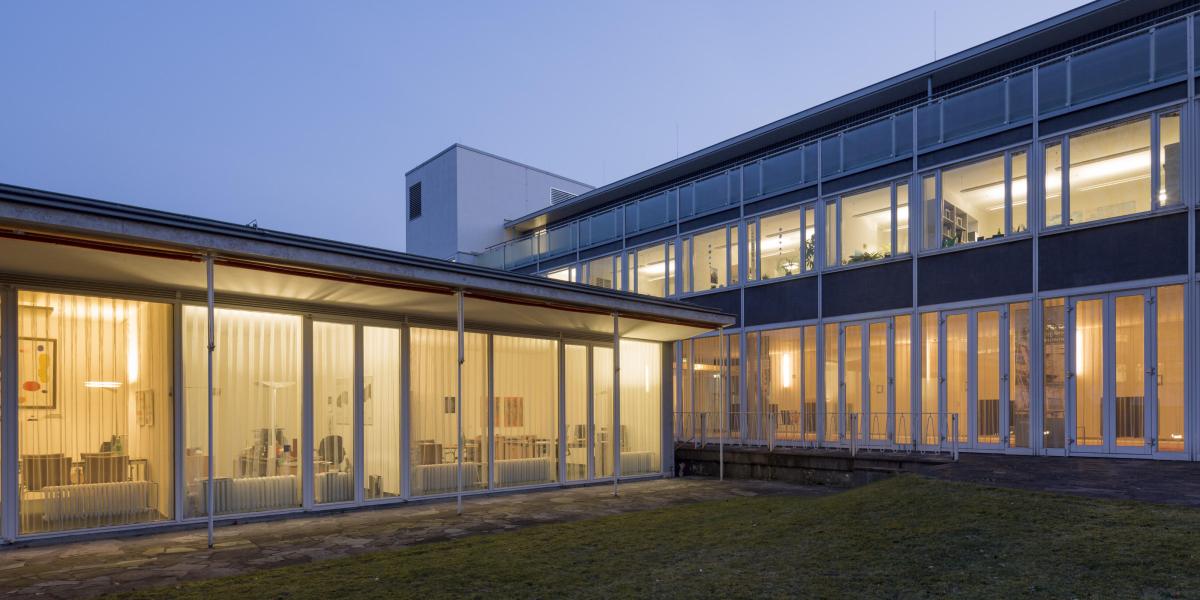 Modernes Bürogebäude mit Glasfassade bei Abenddämmerung, Innenräume warm beleuchtet und transparent einsehbar.