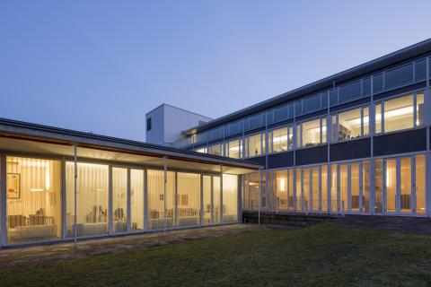 Modernes Bürogebäude mit Glasfassade bei Abenddämmerung, Innenräume warm beleuchtet und transparent einsehbar.