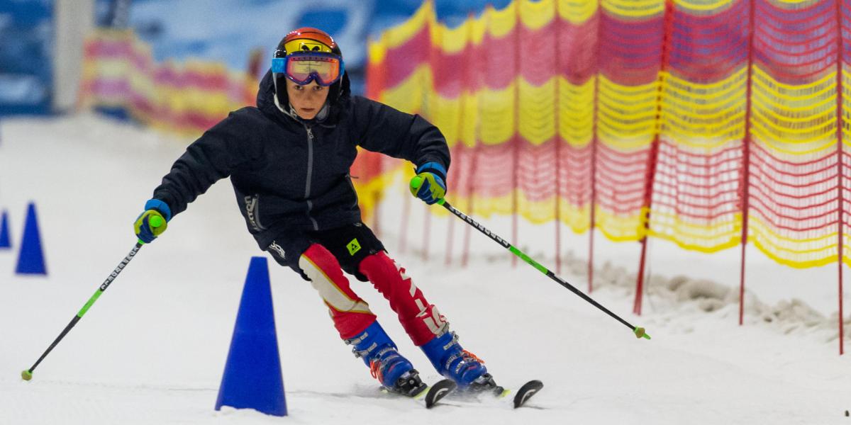 Kind fährt Slalom durch blaue Hütchen auf einer Indoor-Skipiste, geschützt durch bunte Fangnetze, mit vollem Fokus auf der Strecke.