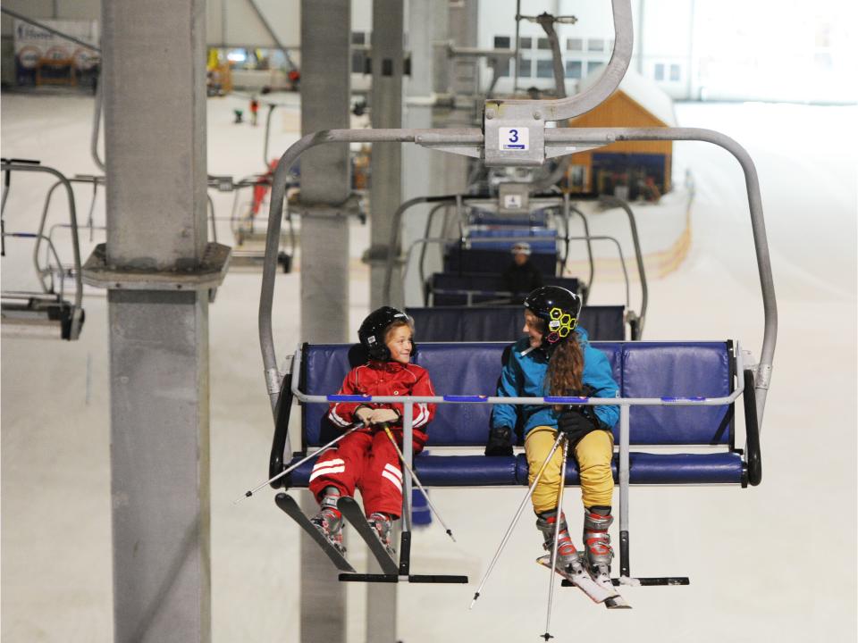 Zwei Kinder in Skikleidung sitzen lachend auf einem Sessellift in einer Indoor-Skihalle mit weitläufiger Schneelandschaft.____