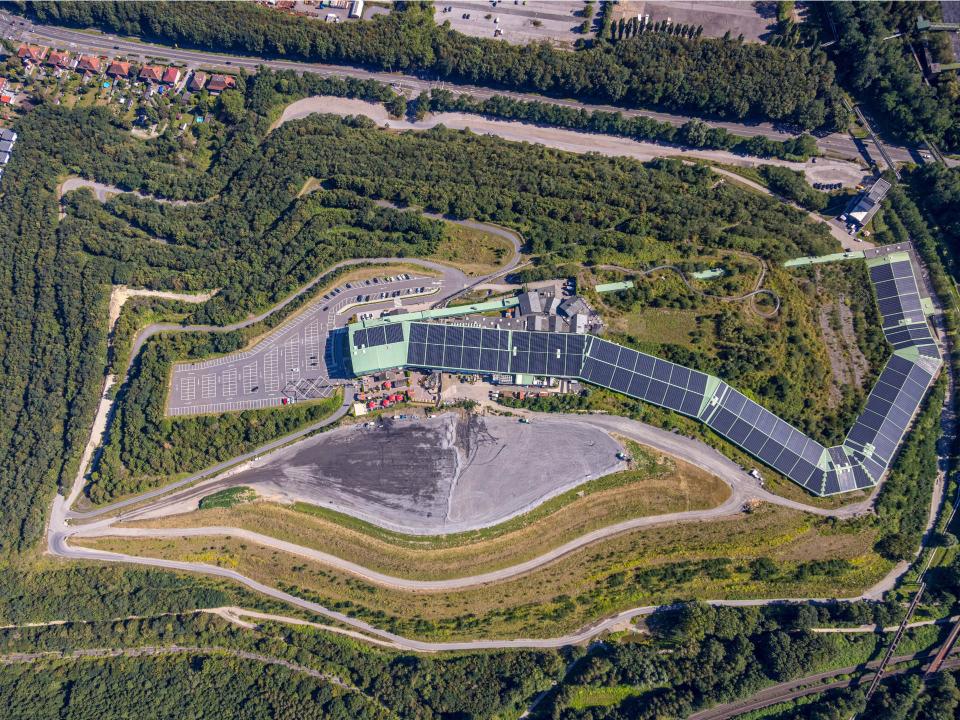 Luftaufnahme einer Skihalle mit Solarpaneelen, umgeben von dichtem Grün und Parkplätzen, integriert in die Landschaft.____