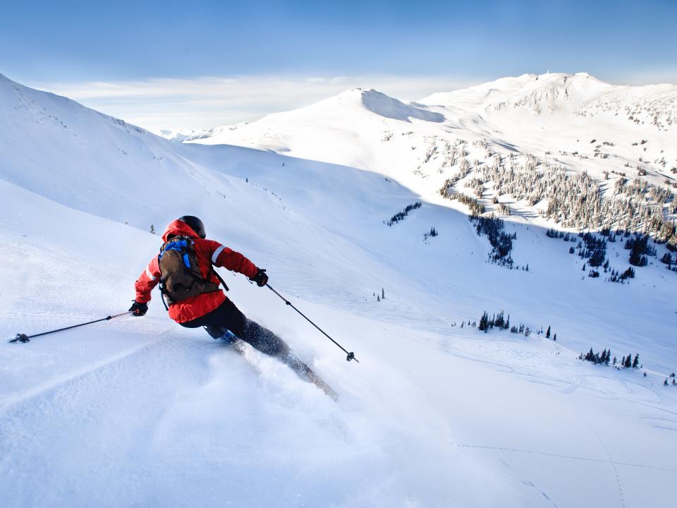 Skifahrer in roter Jacke fährt eine verschneite, unberührte Berghanglandschaft mit atemberaubendem Panoramablick hinab.____