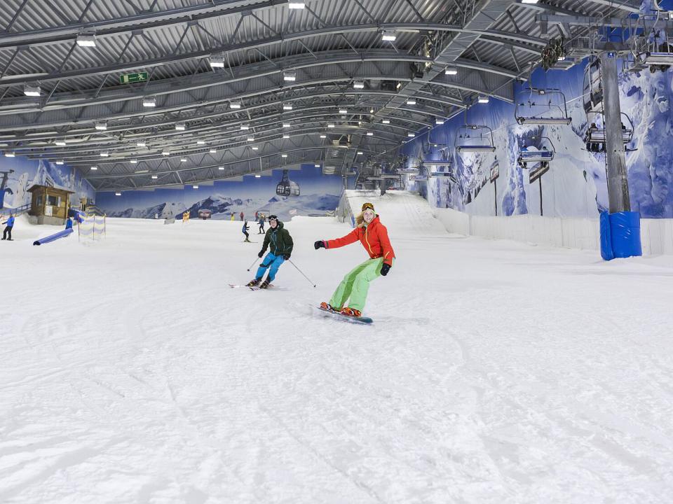Indoor-Skipiste mit Skifahrern und Snowboardern, umgeben von Wandgrafiken alpiner Landschaften, unter einer hohen Hallendecke.____