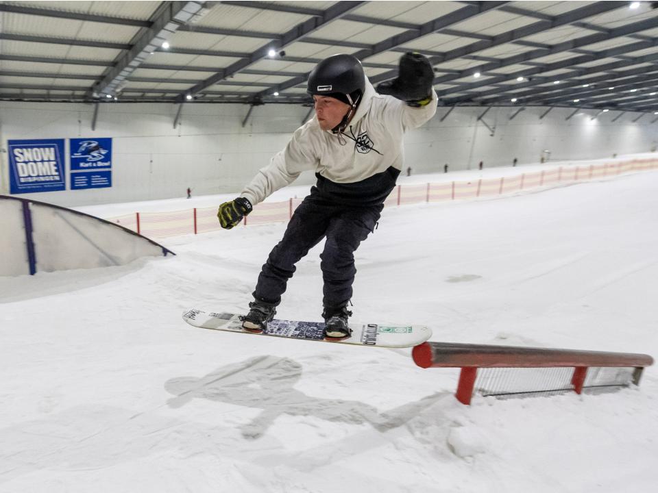 Snowboarder in Sprungpose auf Indoor-Skipiste, balanciert auf einem Rail, in einer Halle mit künstlichem Schnee.____