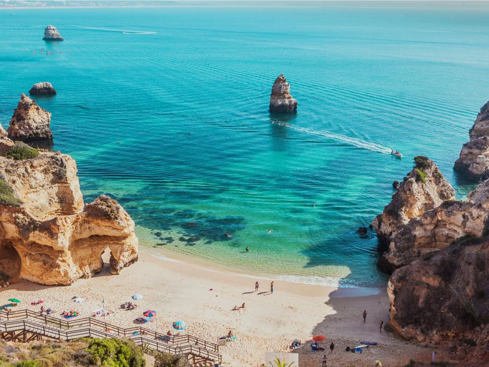 Malerischer Strand mit türkisblauem Wasser, umgeben von Felsen und Badegästen, Boot fährt vorbei.____