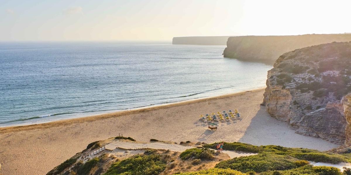 Weitläufiger Sandstrand an einer Küste mit ruhigen Wellen und felsigen Klippen im Hintergrund bei Sonnenuntergang.