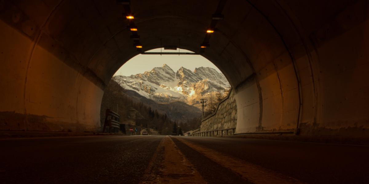 Blick aus einem beleuchteten Straßentunnel auf eine schneebedeckte Berglandschaft bei Sonnenuntergang.