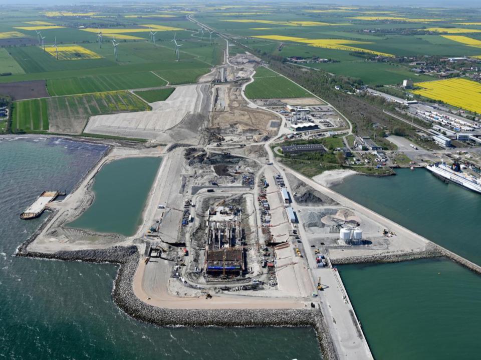 Luftaufnahme einer großen Baustelle am Meer mit angrenzenden Feldern und Windkraftanlagen im Hintergrund.____