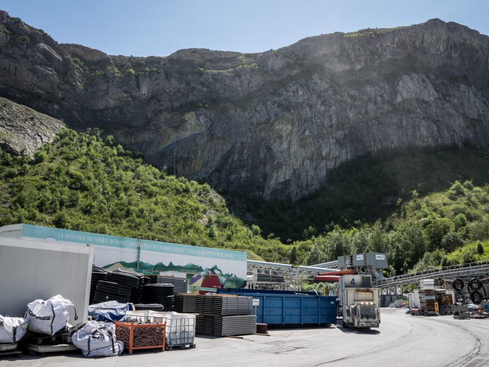 Baustelle mit Materialstapeln und Maschinen vor einer felsigen Berglandschaft im Hintergrund.____