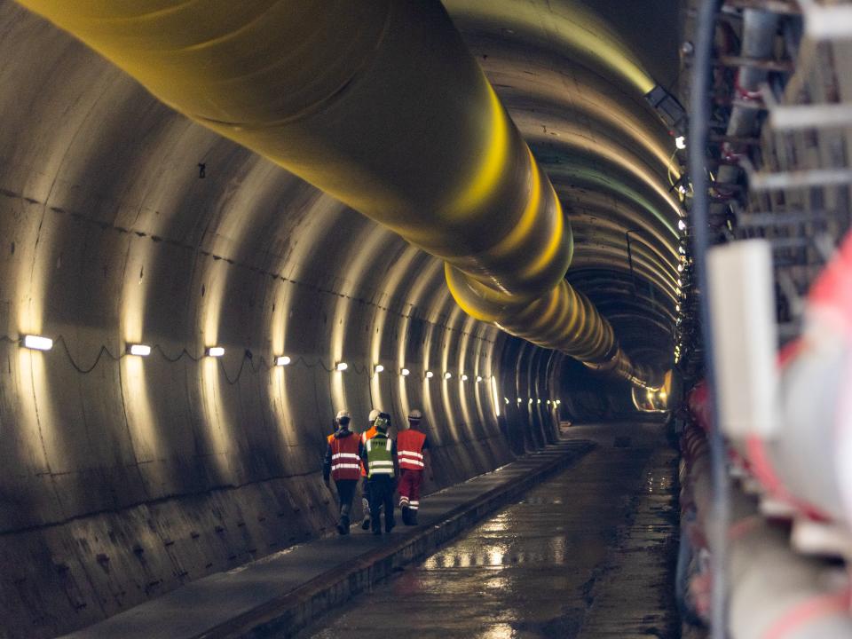 Arbeiter in Warnwesten gehen durch einen beleuchteten Tunnel mit großen Lüftungsrohren.____