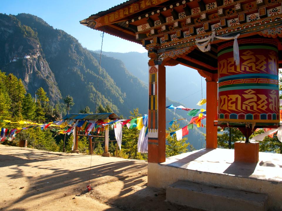 Große Gebetsmühle in Bhutan, umgeben von bunten Gebetsfahnen, mit Berglandschaft im Hintergrund.____