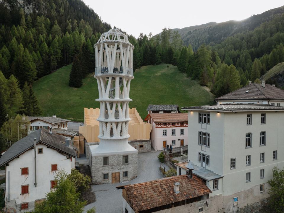 Moderner, weißer Aussichtsturm im Bergdorf mit umliegenden Gebäuden und grünem Berghang im Hintergrund____