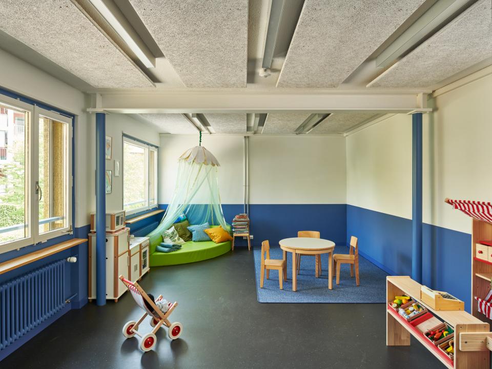 Bunter Kindergartenraum mit Spielzeug, Tisch und Sitzkissen.____