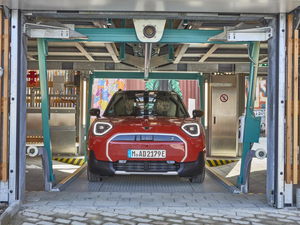Roter E-Mini fährt in automatisierte vertikale Parkanlage mit sichtbarer Stahlstruktur ein.____
