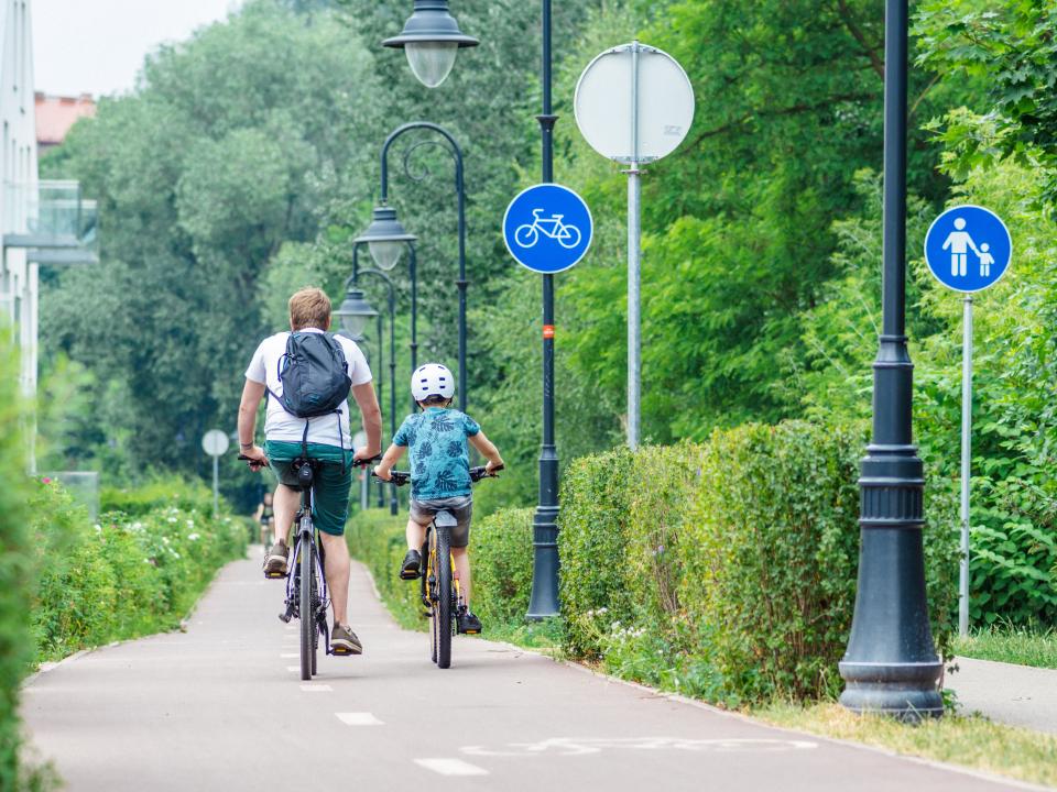Vater und Kind fahren auf einem Radweg in Vilnius, flankiert von Büschen und Verkehrsschildern.____