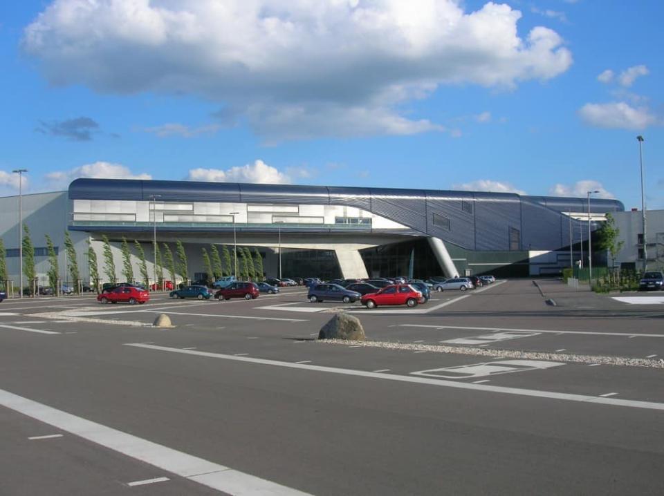 BMW-Werk in Leipzig an einem sonnigen, leicht bewölkten Tag, Vor dem Werk befinden sich ein Parkplatz mit einigen grünenden Bäumen____