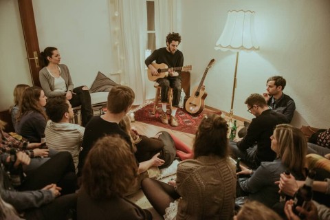 Junge Menschen in einem Wohnzimmer. Ein bärtiger junger Mann spielt gerade Gitarre, die anderen hören gespannt zu.