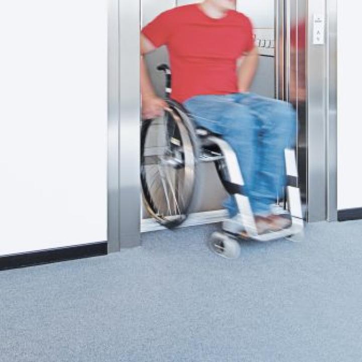 Mann mit rotem T-Shirt und blauer Jeans fährt in seinem Rollstuhl aus einem Fahrstuhl____
