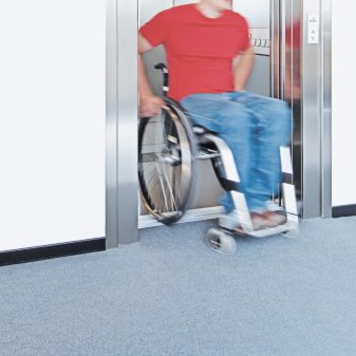 Mann mit rotem T-Shirt und blauer Jeans fährt in seinem Rollstuhl aus einem Fahrstuhl____