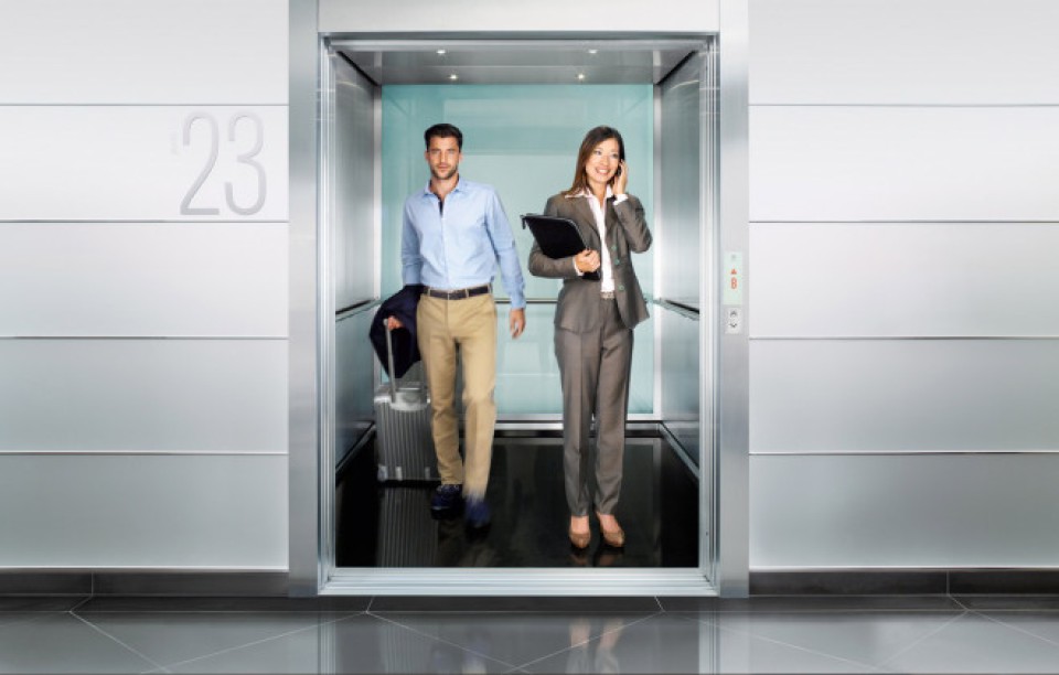 Ein Mann und eine Frau in Anzug stehen in einem Fahrstuhl. Der Mann hat einen Rollkoffer und ist dabei den Fahrstuhl zu verlassen. Die Frau telefoniert lächelnd.____