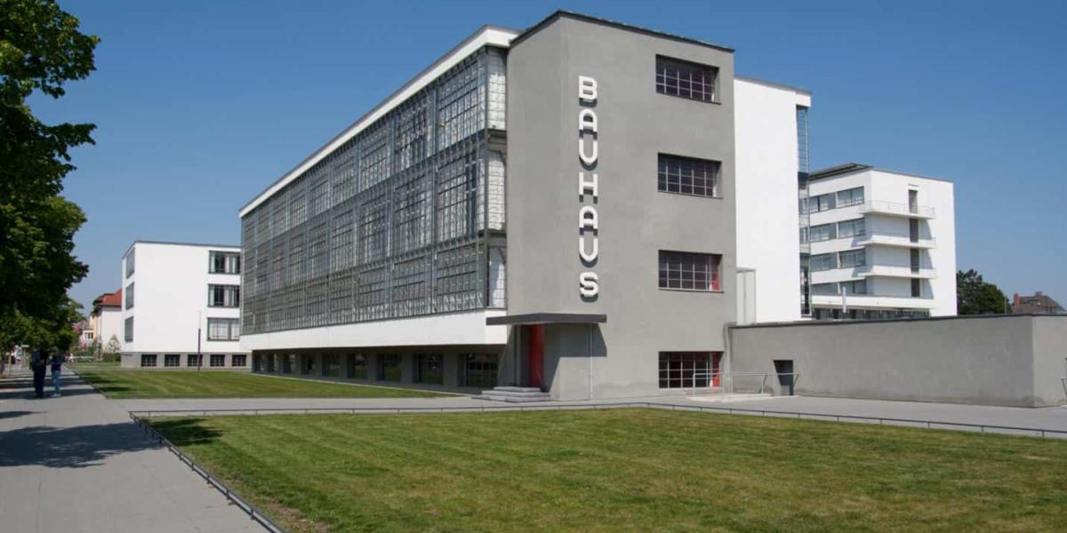 Gebäude der heutigen Bauhaus. Das Schild ist in weißen Initialen, die Wand ist grau, dennoch gibt es viele Fenster und einzelne rot-gefärbte Akzente.