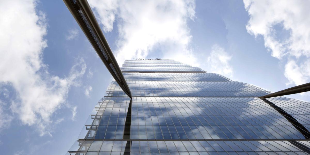 Allianz Tower aus der Froschperspektive. Der Himmel ist klar mit erinnerten Wolken. Die Fassade des Hochhauses ist aus Glas, an ihr ragen zwei Stahlträger.