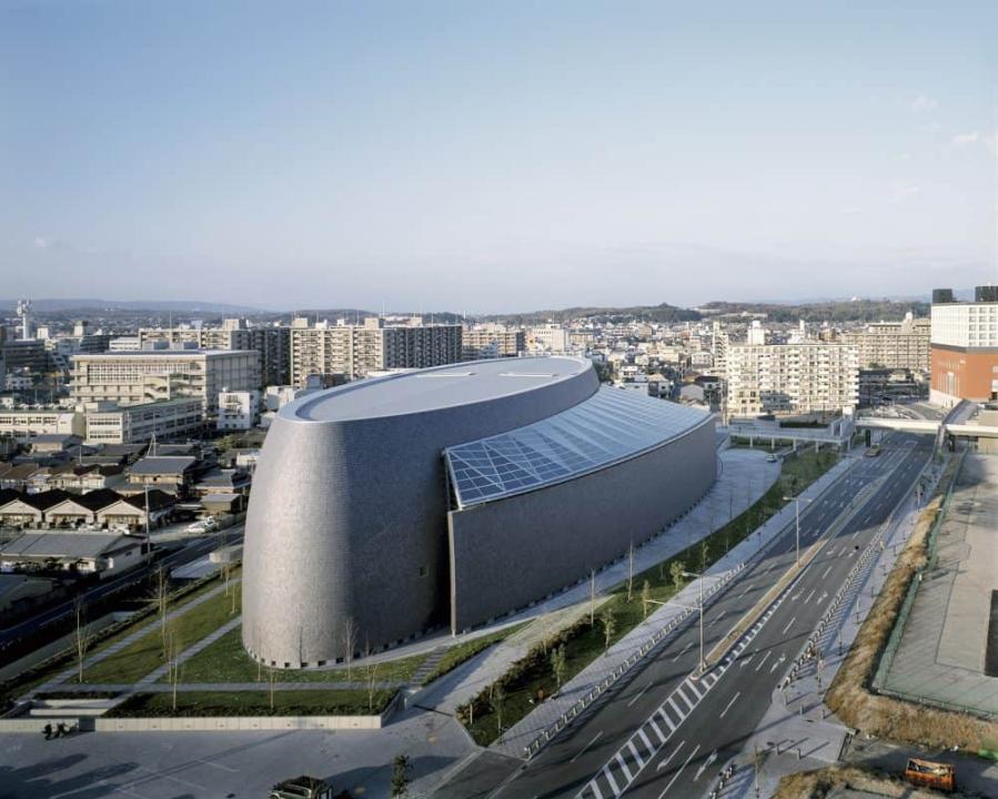Nara Centennial Hall in Japan. Die Halle sieht aus wie ein großer, runder, grauer Block aus, ohne Fenster. Nur der Vorbau ist mit Glas überdacht.____