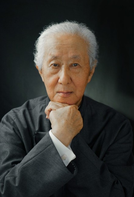 Porter des Architekten Arata Isozaki. er trägt weißes Haar, seine Hände sind vorne ineinander verschränkt. Er trägt einen schwarzen umhäng und ein weißes Hemd.